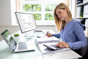Young businesswoman sitting at her desk and using a computer and laptop to calculate bills into a binder.